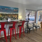 A high-top wooden counter with red metal stools, situated under a large piece of colorful wall art in a brightly lit room